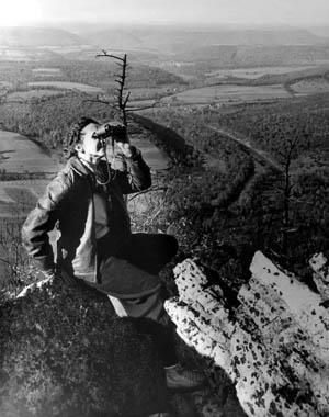 black and white picture of Rachel Carson looking through binoculars on top of a rock with wilderness behind her.
